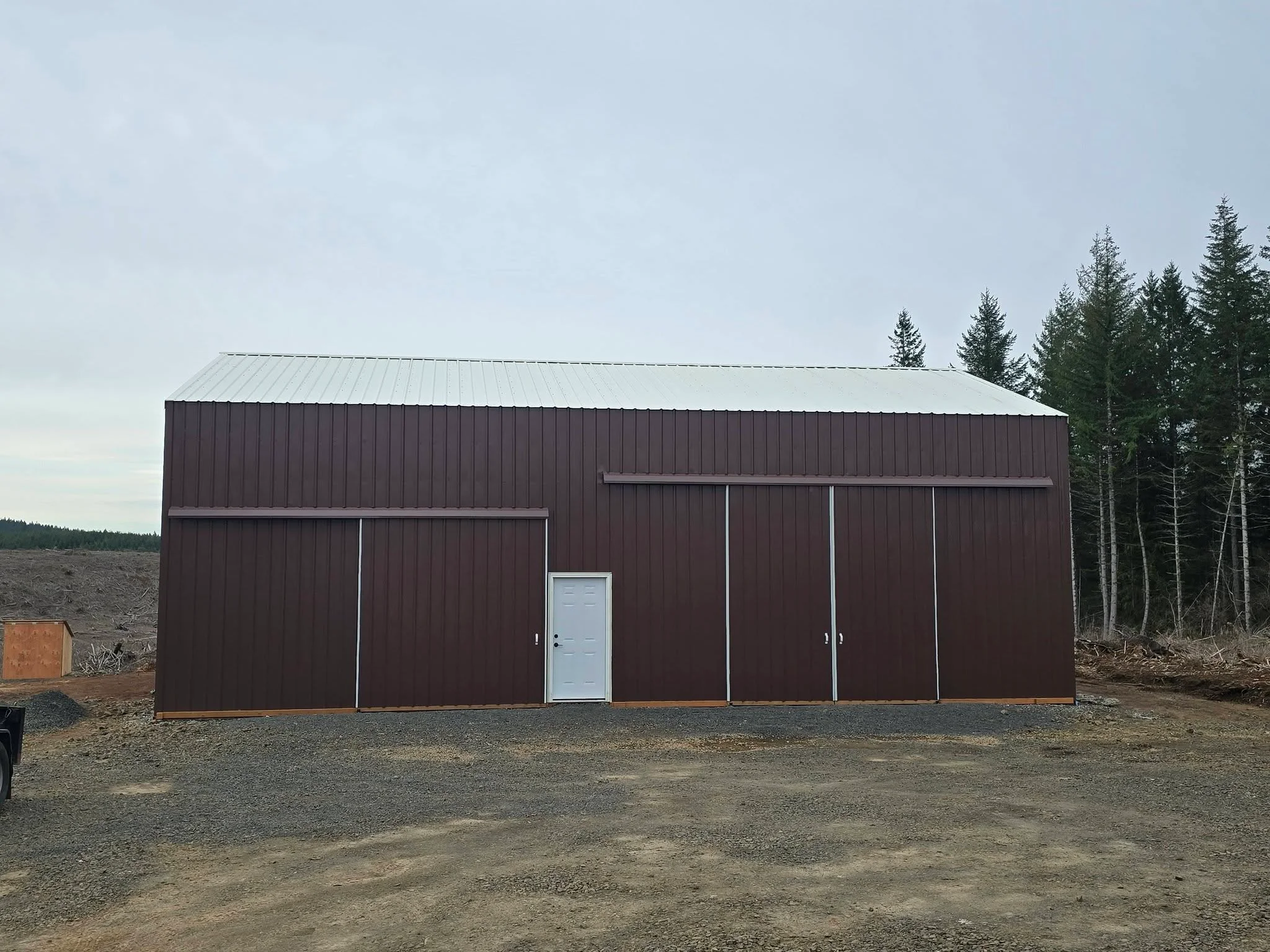 Dark red metal barn with gray roof, white door, and multiple bay openings on gravel foundation
