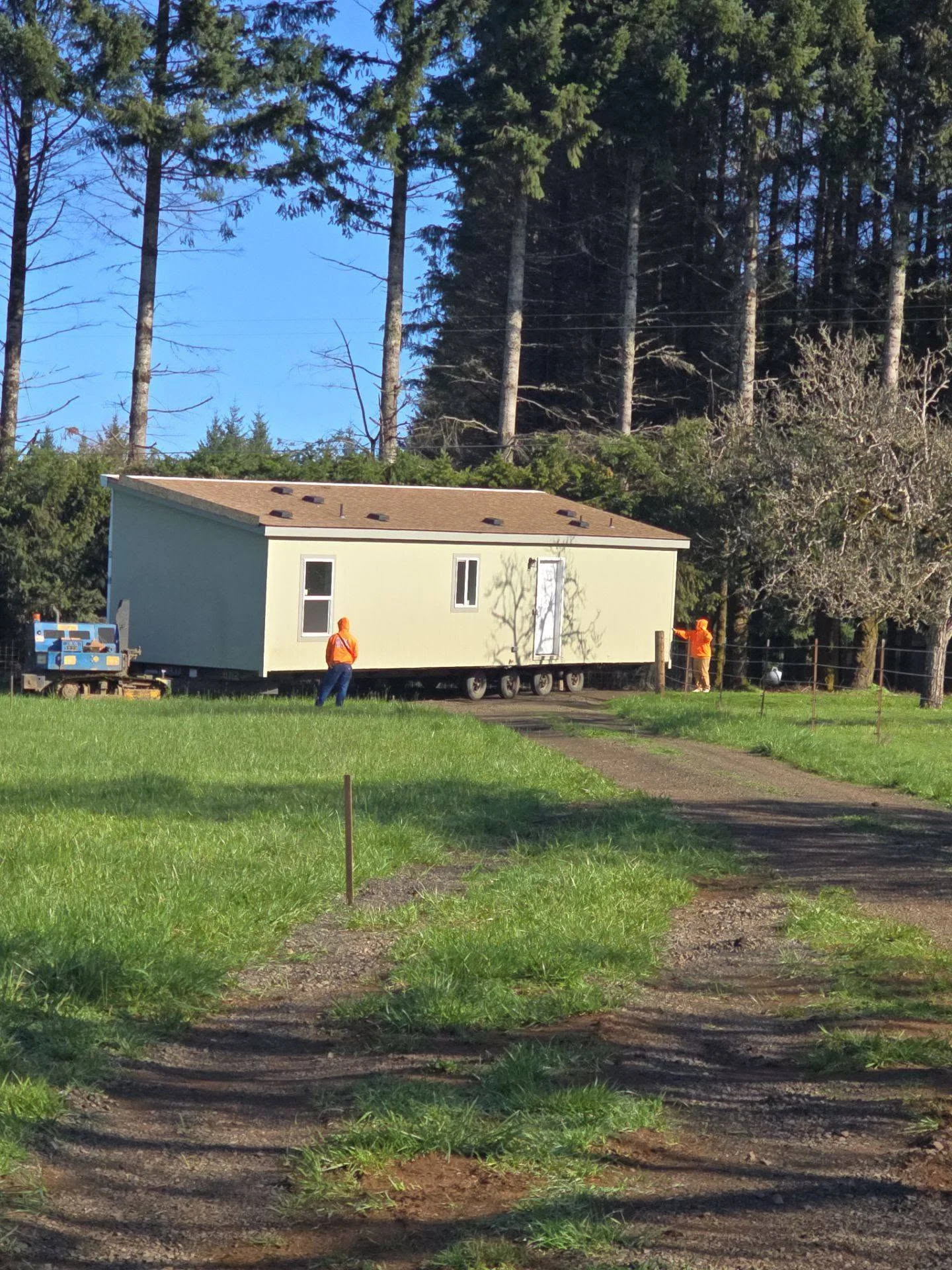 Mobile home installation on gravel lot with workers and forest backdrop