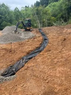 Excavator on sloped dirt site with black drainage fabric laid along contour